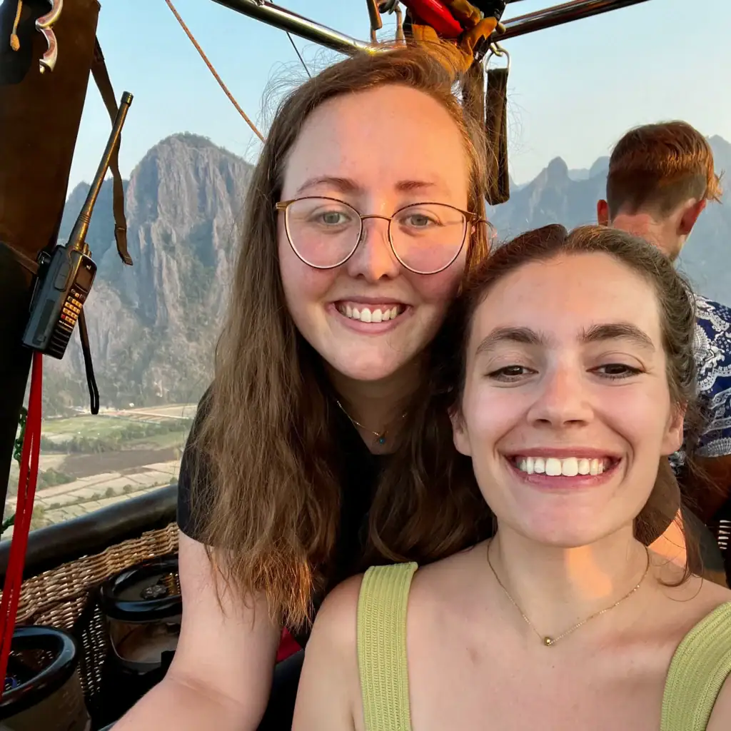 About Us 3 Two woman smiling while standing in the basket of a hot air ballon with the mountains of Vang Vieng, Laos in the background.