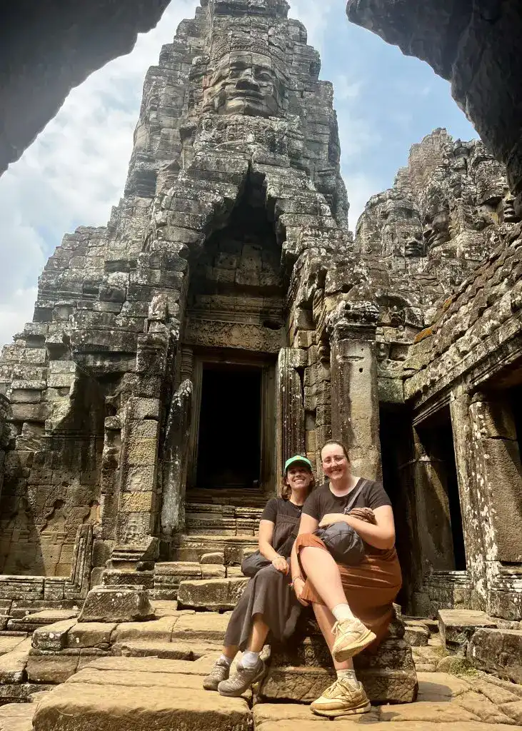About Us 2 Two women posing closely together sitting down in front of a stone tower with a face carved into it at Bayon Temple in Siem Reap, Cambodia.