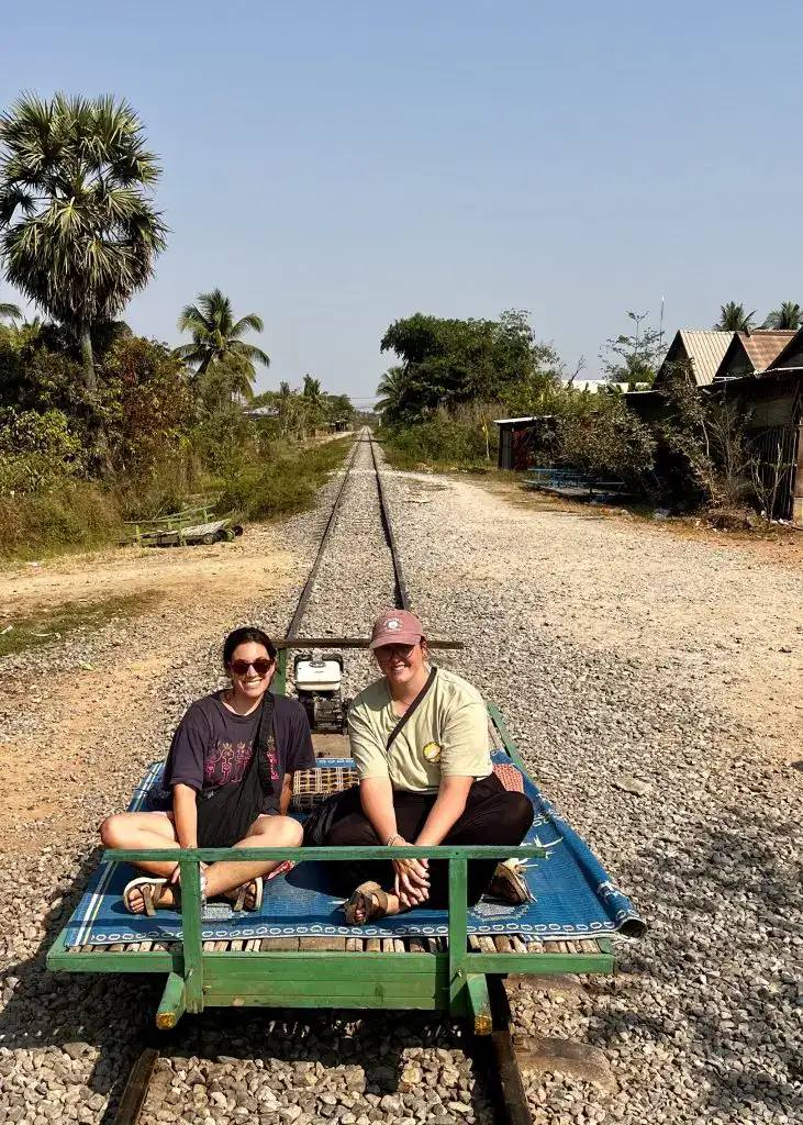 About Us 4 Two women sitting cross legged on the bamboo train located in Battambang, Cambodia.