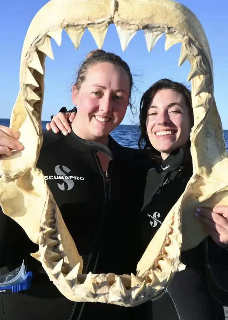 About Us 1 Two women standing on a boat with the ocean in the background with their arms around each other wearing thick wetsuits posing with the jaws of a Great White shark in Port Lincoln, South Australia.